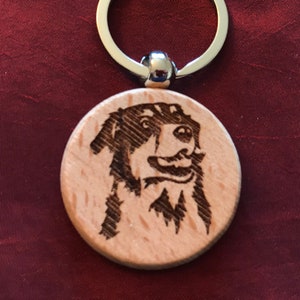 May include: A wooden keychain with a laser-engraved image of a dog's face. The dog has black fur and is looking directly at the viewer. The keychain is attached to a silver metal ring.