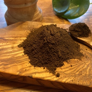 May include: A close-up shot of a pile of dark brown powder on a wooden serving board. A small wooden spoon holds a portion of the powder. The board has a natural wood grain pattern. A wooden grinder is in the background.