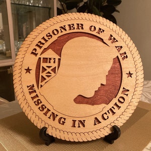 May include: A wooden plaque with a rope border and the text "Prisoner of War Missing in Action" in a red font. The plaque features a silhouette of a person's head and a stylized image of a guard tower.