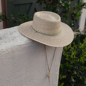 May include: A light brown straw hat with a green ribbon and a wooden bead hanging from the brim. The hat is hanging on a white wall.