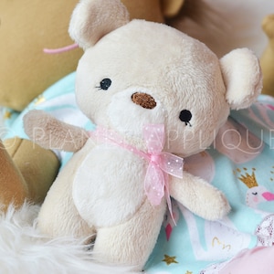 Könnte beinhalten: Ein cremefarbener Plüschteddybär mit einem rosa Band um den Hals. Der Bär sitzt auf einem blauen und weißen gemusterten Stoff mit goldenen Sternen.