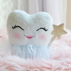 Puede incluir: Un diente de peluche blanco con una cara sonriente, que lleva una falda de tul azul y sostiene una varita dorada con forma de estrella. El diente está sentado sobre una superficie rosa y esponjosa.