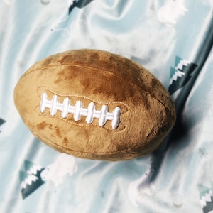 Puede incluir: Una pelota de fútbol de peluche marrón con costuras blancas. La pelota está sobre una tela de estampado azul y blanco.