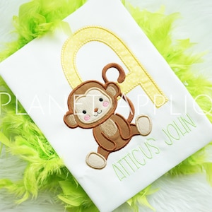 May include: A white t-shirt with a yellow letter A with white polka dots. A brown monkey with a pink nose and cheeks is hanging from the letter A. The name "Atticus John" is embroidered in green below the monkey.