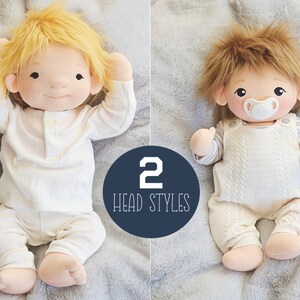 Op de afbeelding: Twee handgemaakte knuffels met verschillende kopvormen. De pop links heeft kort, glad, blond haar en draagt een witte romper met lange mouwen. De pop rechts heeft lang, bruin, ruig haar en draagt een witte romper met een speen in zijn mond. De tekst "2 HEAD STYLES" staat in een blauwe cirkel.