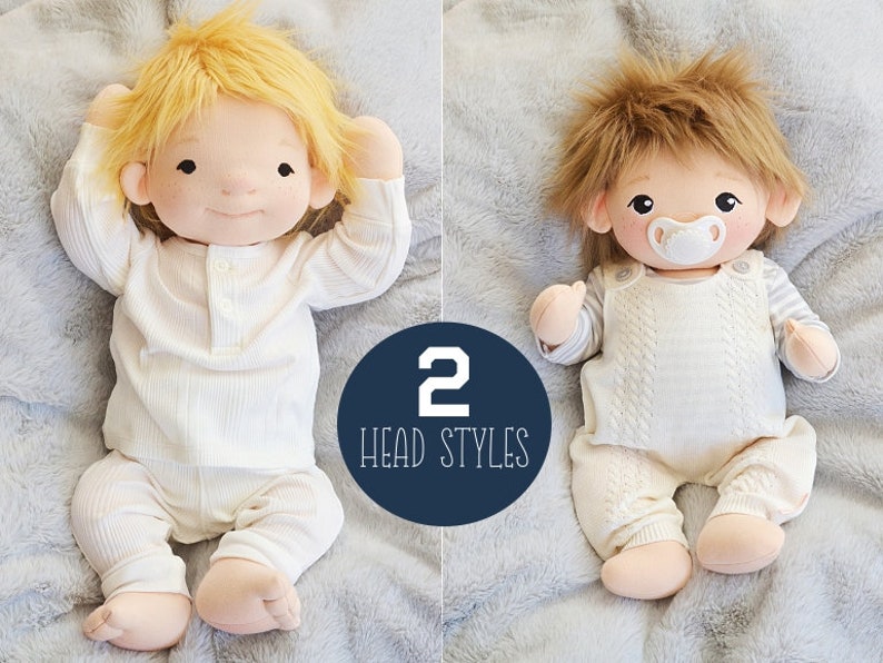 Op de afbeelding: Twee handgemaakte knuffels met verschillende kopvormen. De pop links heeft kort, glad, blond haar en draagt een witte romper met lange mouwen. De pop rechts heeft lang, bruin, ruig haar en draagt een witte romper met een speen in zijn mond. De tekst "2 HEAD STYLES" staat in een blauwe cirkel.