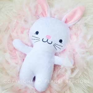 May include: A white plush bunny with pink inner ears, a pink nose, and embroidered black eyes and whiskers. The bunny is lying on a bed of pink feathers. The text "COPYRIGHT © PLANET APPLIQUE" is visible.
