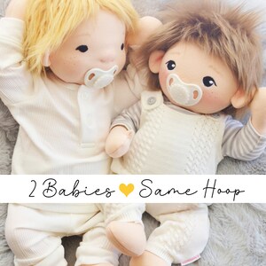 Op de afbeelding: Twee handgemaakte babypoppen van stof met speen, die witte outfits dragen, liggen op een pluche grijs oppervlak. De poppen hebben blond en bruin haar. De tekst "2 Babies &hearts; Same Hoop" is onder de poppen gedrukt.