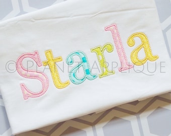 May include: A white fabric with the name "Starla" embroidered in colorful letters. The letters are outlined in white and filled with different patterns, including polka dots, stripes, and checkered.