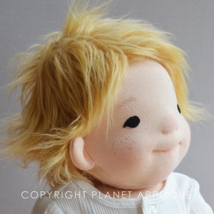Op de afbeelding: Een close-up van een handgemaakte pop met blond haar en een wit shirt. De pop heeft een geschilderd gezicht met gesloten ogen en een kleine, licht geopende mond. Het haar van de pop is gestyled in een rommelige, warrige look.