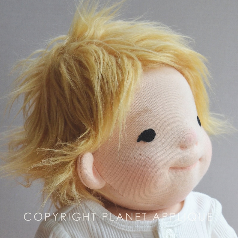 Op de afbeelding: Een close-up van een handgemaakte pop met blond haar en een wit shirt. De pop heeft een geschilderd gezicht met gesloten ogen en een kleine, licht geopende mond. Het haar van de pop is gestyled in een rommelige, warrige look.
