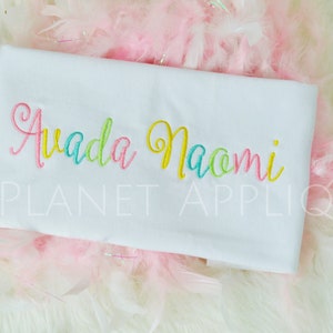 May include: White fabric with the names "Awada Naomi" embroidered in colorful cursive lettering. The text is in a rainbow color scheme with yellow, green, pink, and blue.