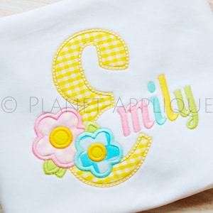 May include: A yellow gingham letter E with a pink, yellow, and blue flower design. The name "Emily" is embroidered in pink, yellow, and blue.