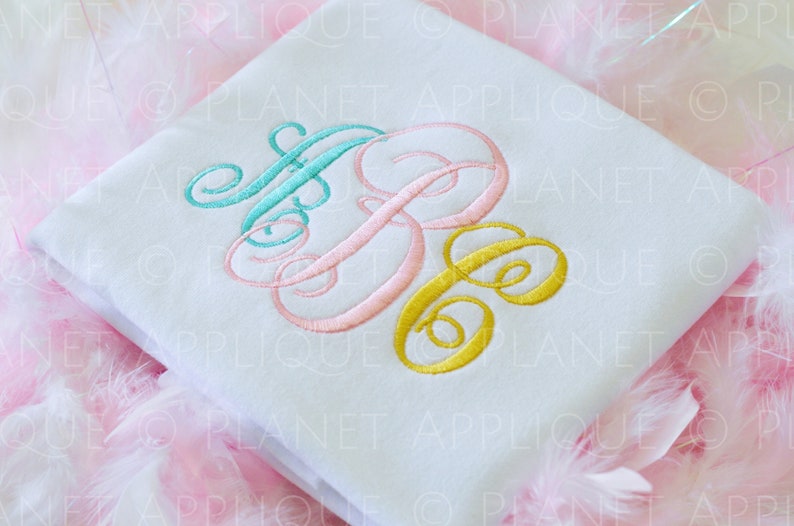 May include: A white t-shirt with a monogram embroidered in turquoise, pink, and gold thread. The monogram is a stylized "ABC" with the letters intertwined.