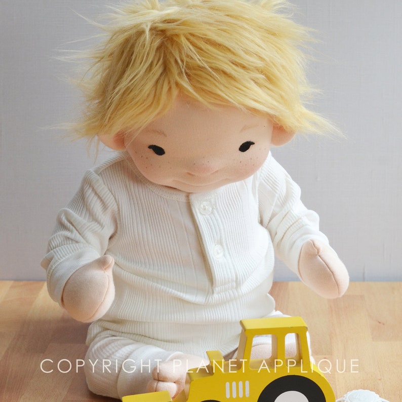 Op de afbeelding: Een handgemaakte pop met blond haar die een wit shirt met lange mouwen draagt en met een gele speelgoedtruck speelt. COPYRIGHT PLANET APPLIQUE is gedrukt op de houten vloer.