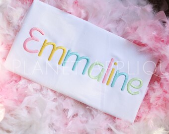 Op de afbeelding: Witte stof met de naam "Emmaline" geborduurd in kleurrijke letters. De letters zijn omrand met geel, roze, blauw en groen.