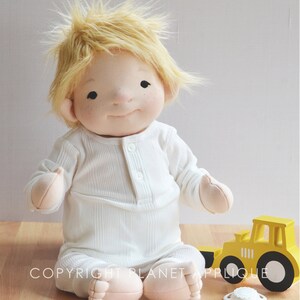 Op de afbeelding: Een handgemaakte stoffen pop met blond haar die een wit geribbeld rompertje draagt. De pop zit op een houten oppervlak met een gele speelgoedtruck en een speen.