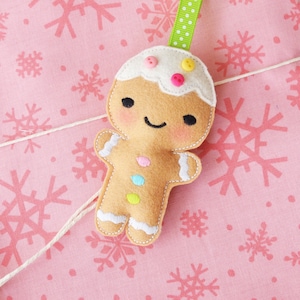May include: A gingerbread man ornament with a white icing top and colorful buttons. The ornament is hanging from a green and white ribbon against a pink background with snowflake patterns.