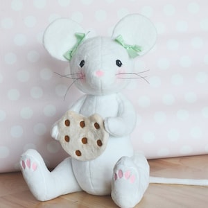 May include: White plush mouse toy with green bows, holding a brown chocolate chip cookie. The mouse is sitting on a wooden floor.