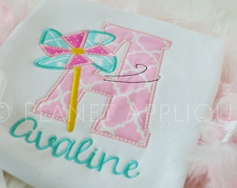 May include: A pink and white patterned letter A with a turquoise and pink pinwheel on a white shirt. The name "Avaline" is embroidered in turquoise below the letter.