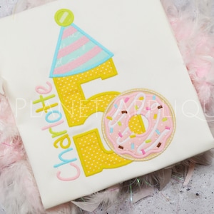 Könnte beinhalten: Ein weißes T-Shirt mit dem Namen "Charlotte" und der Zahl "5" in Gelb mit rosa Punkten gestickt. Neben der Zahl ist ein rosa Donut mit Streuseln gestickt. Die Zahl "5" hat einen Partyhut oben drauf.
