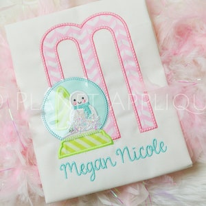 Könnte beinhalten: Ein weißes Shirt mit einem pink-weißen Chevron-Buchstaben M, einer blauen und grünen Schneekugel mit einem Schneemann darin und dem Namen Megan Nicole in blauer Stickerei.