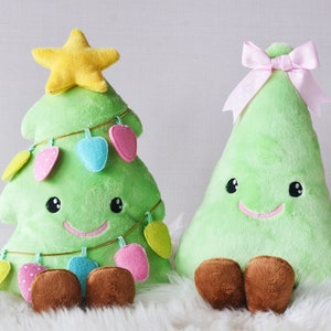 Puede incluir: Dos juguetes de peluche de árbol de Navidad verde con caras sonrientes. Un árbol tiene una estrella amarilla en la parte superior y luces de colores, el otro tiene un lazo rosa.
