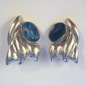May include: A pair of silver-toned earrings featuring abstract, fluid metal shapes with polished surfaces. Each earring is set with an oval-shaped, iridescent abalone shell inlay displaying deep blue and green tones with natural dark veining.