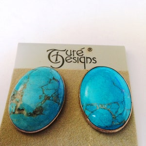 May include: A pair of sterling silver earrings with turquoise stones. The earrings are oval shaped and have a light blue color with dark gray veins. The earrings are on a white card with the text "Turé Designs" and "Sterling Silver".
