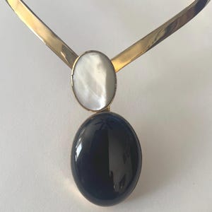 May include: A gold-toned choker necklace with a pendant featuring a white oval stone above a large, dark blue oval stone. The necklace has a simple, elegant design, suitable for formal or casual wear.