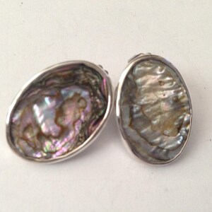 May include: Two oval-shaped earrings with iridescent abalone shells set in silver. The shells display a range of colors, including pink, purple, and green. The earrings are displayed on a white surface.
