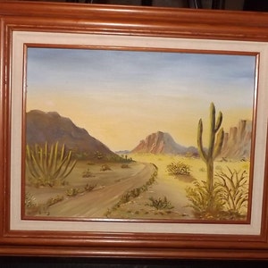 May include: A framed oil painting depicting a desert landscape with a dirt road leading into the distance. The painting features a large cactus in the foreground, with mountains and a blue sky in the background.