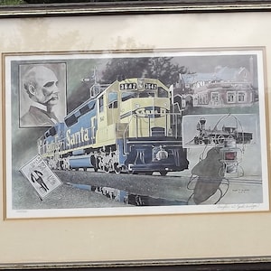 May include: A framed print of a Santa Fe train with a blue and yellow locomotive. The print includes a portrait of a man, a building, and a smaller steam engine. The text "Santa Fe" is visible on the side of the train.