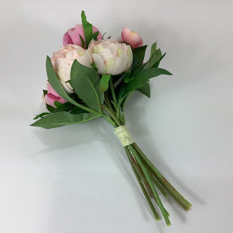 May include: A bouquet of artificial peonies in shades of pink and cream, with green leaves and stems. The flowers are clustered together and tied with a light-colored ribbon.