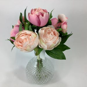 May include: A bouquet of pink and peach peonies with green leaves in a clear, round glass vase. The vase has a diamond pattern. The flowers are in various stages of bloom, with some tightly closed and others fully open.