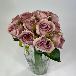 May include: A bunch of mauve artificial roses in a clear, ribbed glass vase. The roses have multiple petals and green leaves. The vase is partially filled with water, and the arrangement is set against a white backdrop.