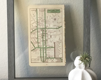 Può includere: Una mappa vintage incorniciata di Dallas, Texas, che mostra il centro degli affari. La mappa è in una cornice grigia con un fronte di vetro. Un vaso di ceramica bianco con una pianta aerea verde si trova su una superficie marrone scuro.