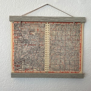 May include: A vintage map of Los Angeles, California, printed on paper and mounted in a wooden frame. The map shows the city's streets and neighborhoods, including Beverly Hills, Hollywood, and Downtown Los Angeles. The map is divided into sections, with each section labeled with a letter and a number. The map is in black and white, with red lines indicating the streets.