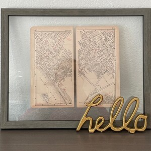 May include: Framed vintage map print with a gray wooden frame. The map is divided into two sections, with red markings and text. A gold-colored "hello" sign is placed in front of the frame.