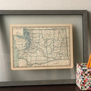 May include: Framed vintage map of Washington state with a gray wooden frame. The map is in shades of beige, blue, and green. A geometric patterned pencil holder and several yellow pencils with red erasers are next to the frame.