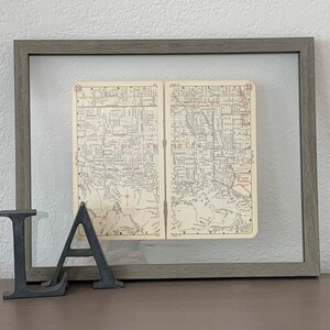 May include: A framed vintage map of Los Angeles, California, with the text "Los Angeles" and "Universal City" visible. The map is in a gray frame and is sitting on a wooden surface with the letters "LA" in front of it.