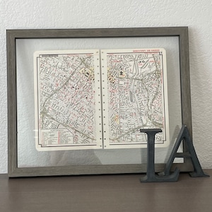 Puede incluir: Un mapa vintage enmarcado del centro de Los Ángeles, California, con un mapa en blanco y negro y texto rojo. El mapa está en un marco gris con un frente de vidrio. El marco está sentado sobre una superficie de madera marrón. Las letras "LA" están frente al marco.