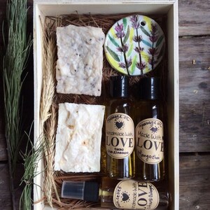 May include: A wooden box filled with bath and body products. The box contains two bars of soap, two bottles of massage oil, and a bottle of spray. The massage oil bottles have labels that say "Handmade with Love" and "Massage Oil". The soap bars are white and have a speckled appearance. The box is lined with straw.
