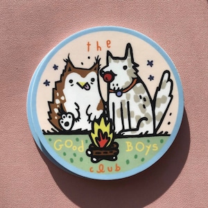 May include: A round sticker with a light blue border and a pink background. The sticker features a cartoon illustration of a dog and a cat sitting by a campfire. The text "the good boys club" is written around the campfire.