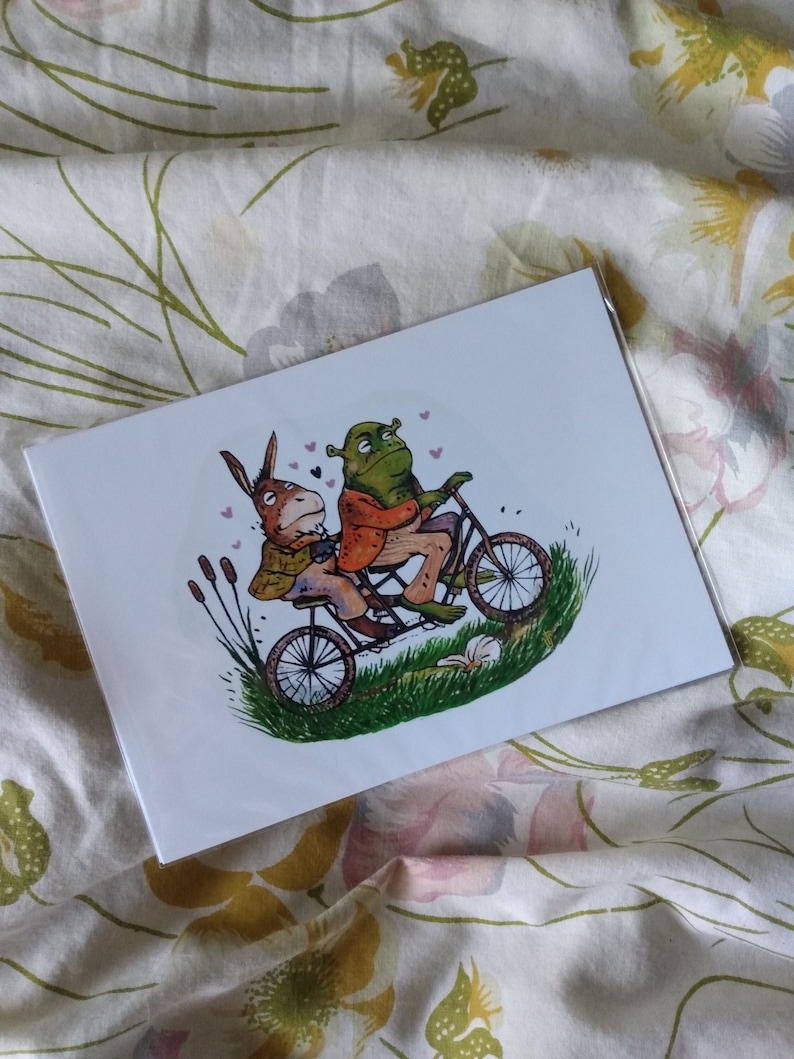May include: A greeting card showing a donkey and a green ogre riding a tandem bicycle. The illustration includes grass, cattails, and small pink hearts. The card is in a clear plastic sleeve, set against a floral background.