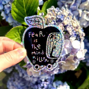 Peut inclure: Un autocollant holographique en forme de pomme avec un centre noir portant le texte "Fear is the mind killer" en lettres blanches. Le motif comprend une illustration stylisée d'un ver à l'intérieur de la pomme et une feuille sur la tige.