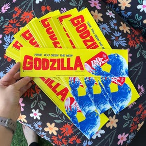 Puede incluir: Una pila de pegatinas rectangulares amarillas con el texto rojo HAVE YOU SEEN THE NEW GODZILLA y una ilustración azul de Godzilla sosteniendo una lata de Dr Pepper roja y blanca. Las pegatinas tienen un fondo amarillo brillante y texto negro.