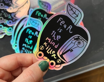 Könnte beinhalten: Ein holografischer Aufkleber mit einem schwarzen Apfel-Design. Der Apfel hat einen weißen Umriss und den Text "Fear is the Mind Killer" im Inneren. Der Aufkleber ist schillernd und hat einen Regenbogeneffekt.