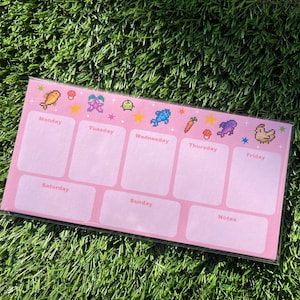 May include: A pink weekly planner with pixelated graphics of a fish, flower, and other icons. The days of the week are labeled, with space for notes. The planner is rectangular and sits on a bed of green grass.