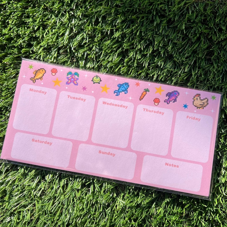 May include: A pink weekly planner with pixelated graphics of a fish, flower, and other icons. The days of the week are labeled, with space for notes. The planner is rectangular and sits on a bed of green grass.